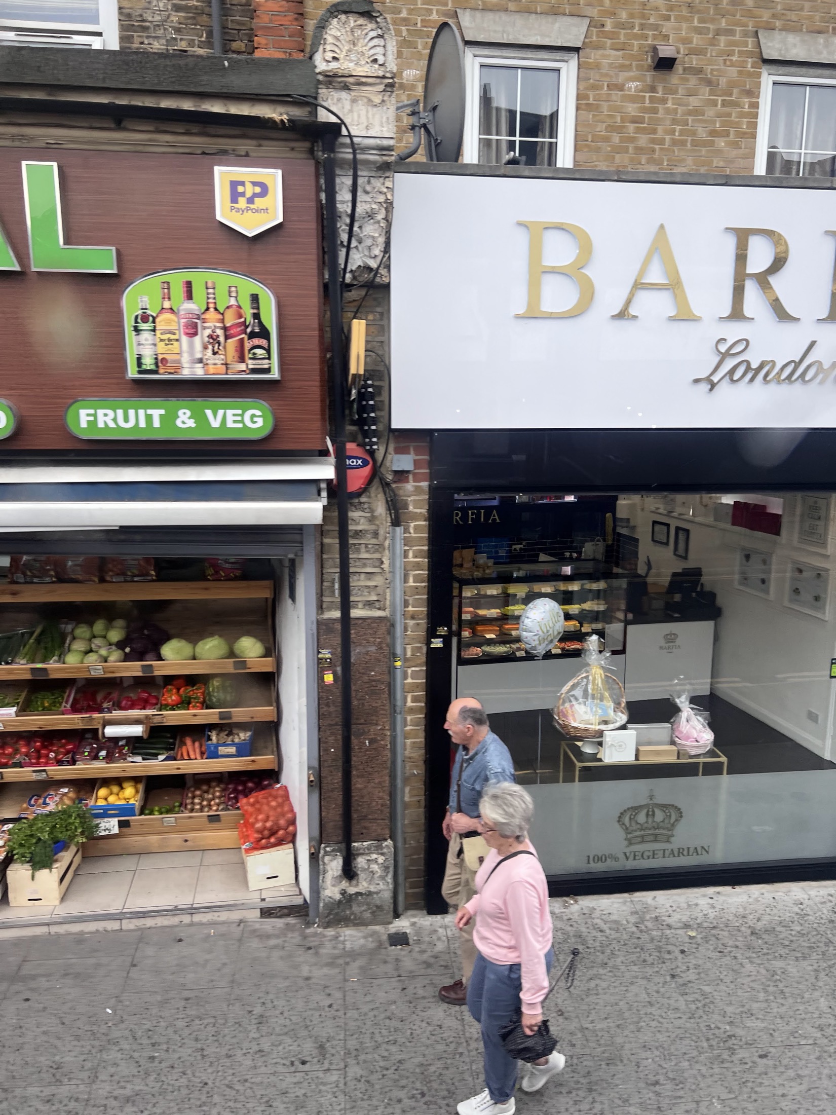 A close view of the Central Wine shopfront, showing the illuminated spirits display, the round Off Licence Open Till Late sign and the fruit and veg stall on the pavement below.