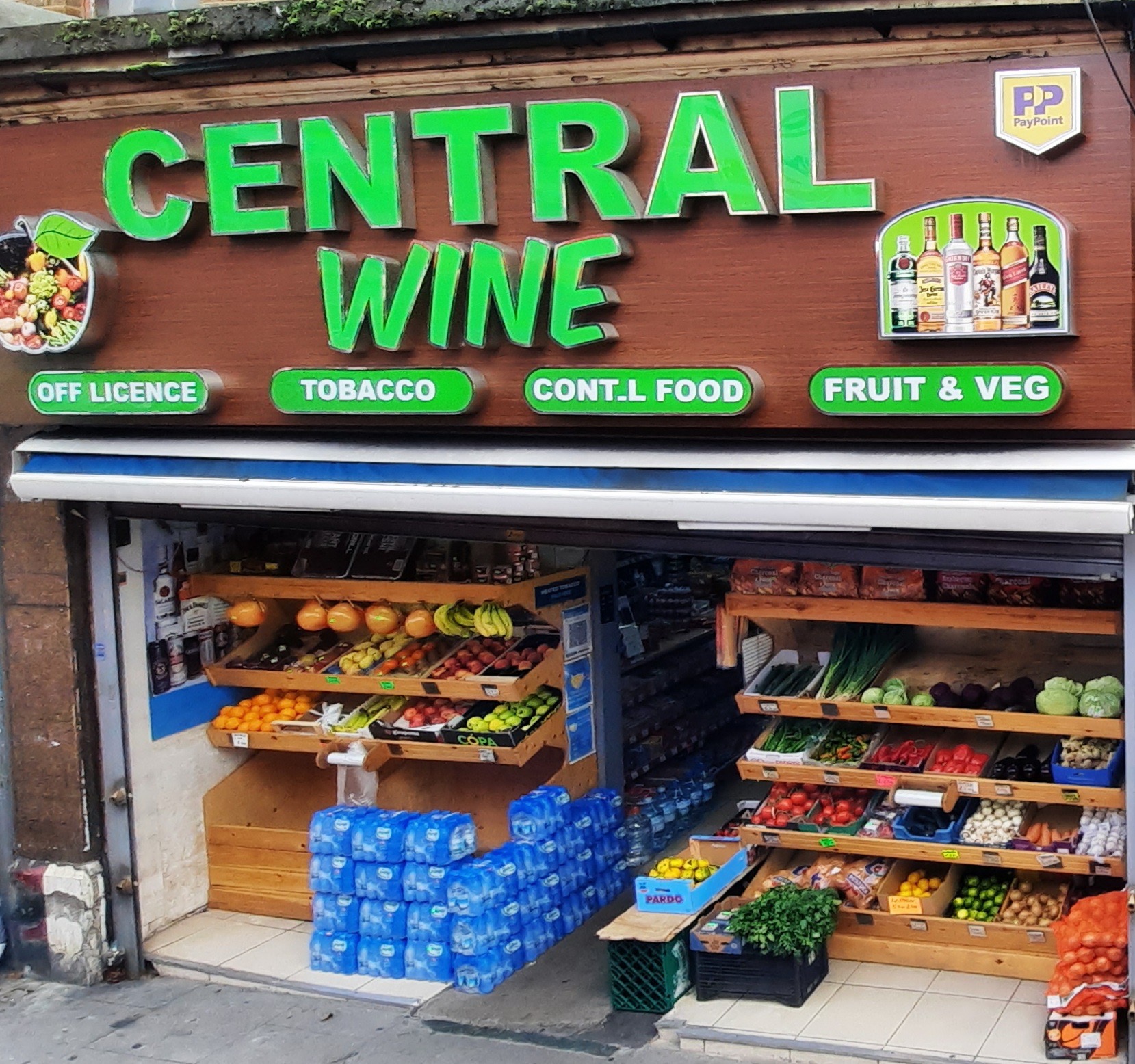 The Central Wine shopfront on Hoe Street, its green sign reading Central Wine above panels for off licence, tobacco, continental food and fruit and veg, with a display of spirits and a fruit and veg stall below.